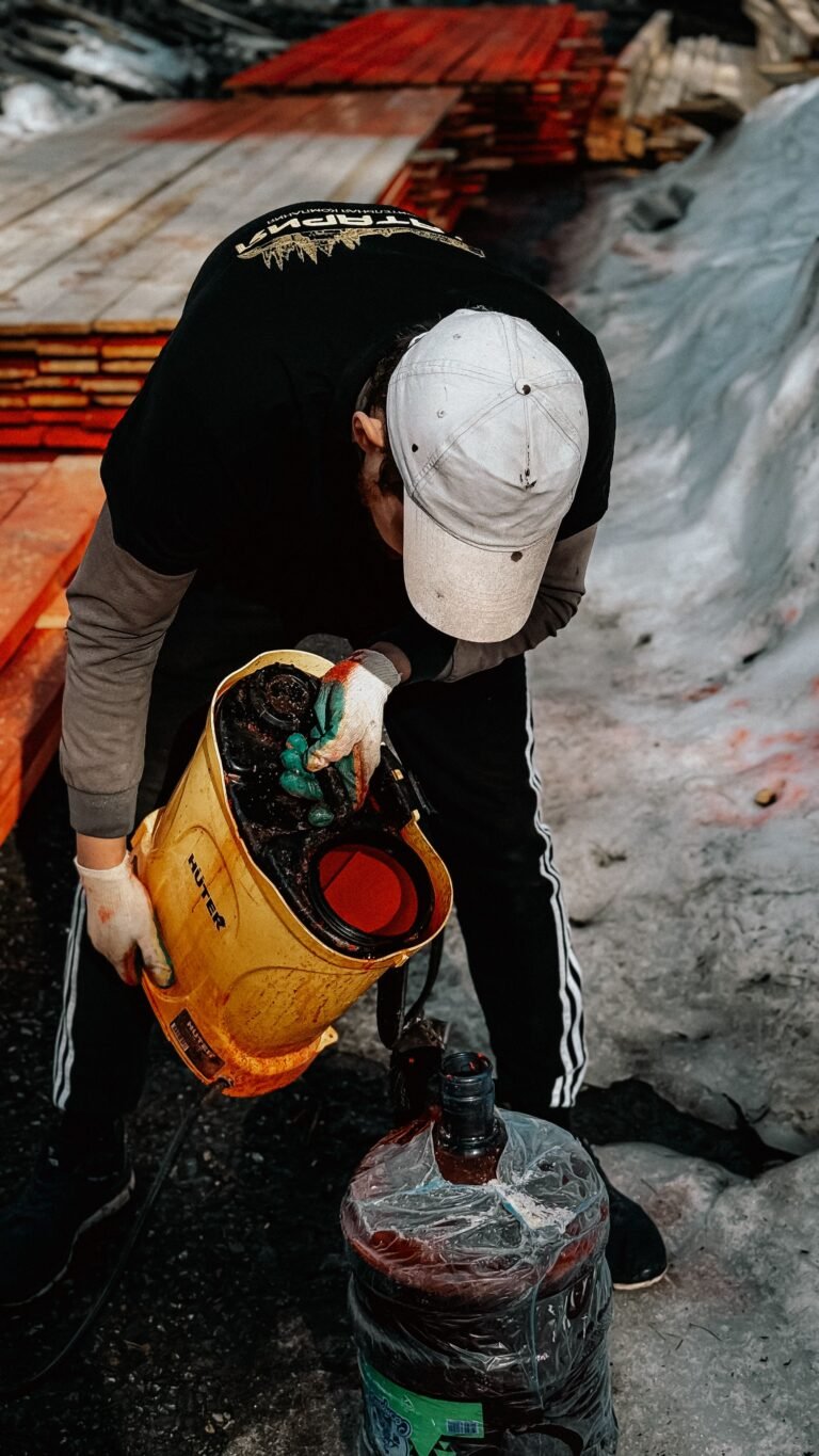 A worker applying protective stain to lumber outdoors during day using a sprayer, preparing for construction.