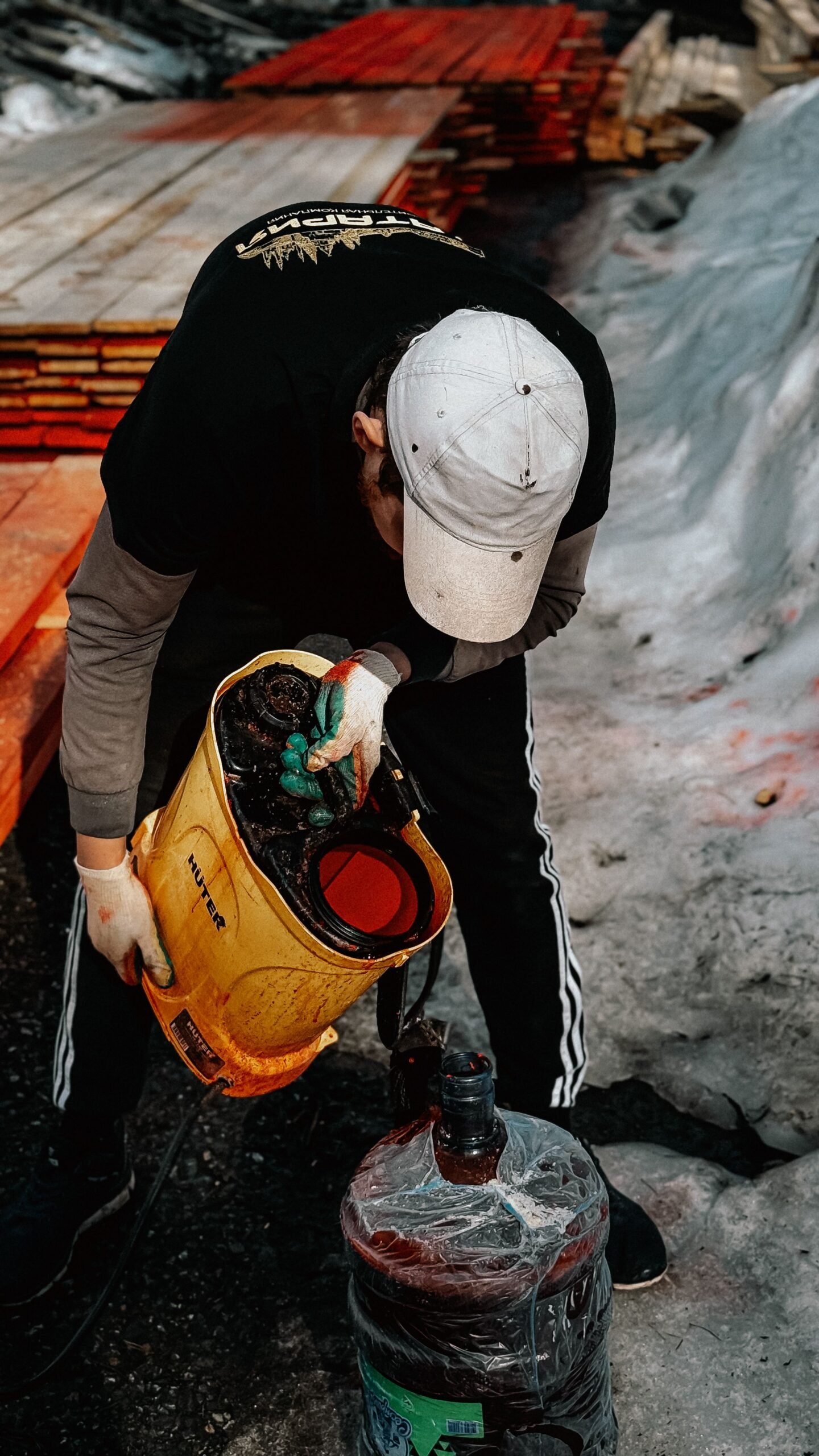 A worker applying protective stain to lumber outdoors during day using a sprayer, preparing for construction.