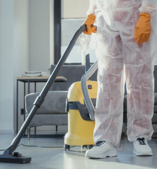 A cleaner in protective coveralls and gloves vacuuming a modern living room.